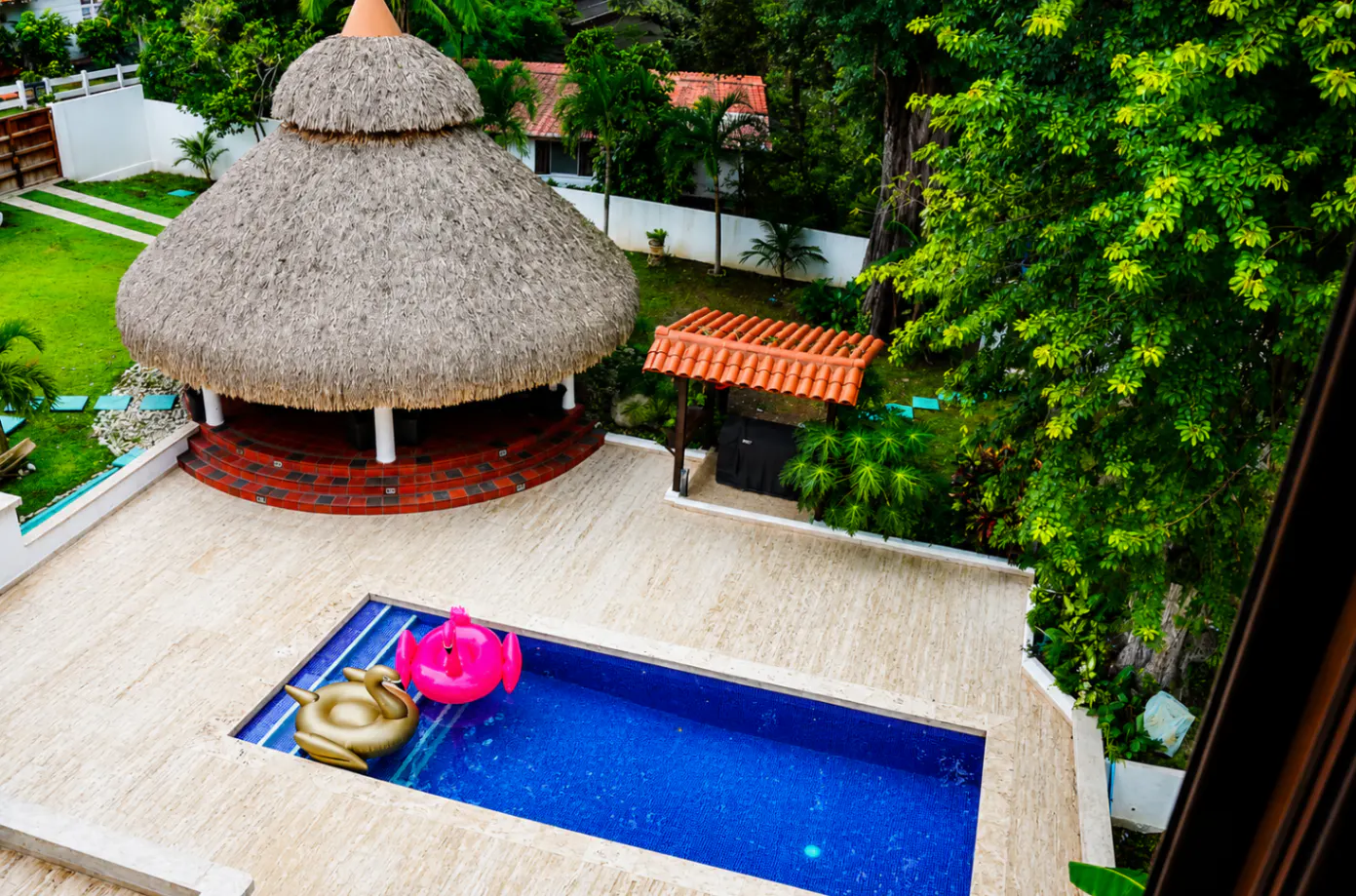 Aerial view of a backyard with a blue pool, pink flamingo and gold swan floats, a thatched-roof gazebo, and lush green trees.