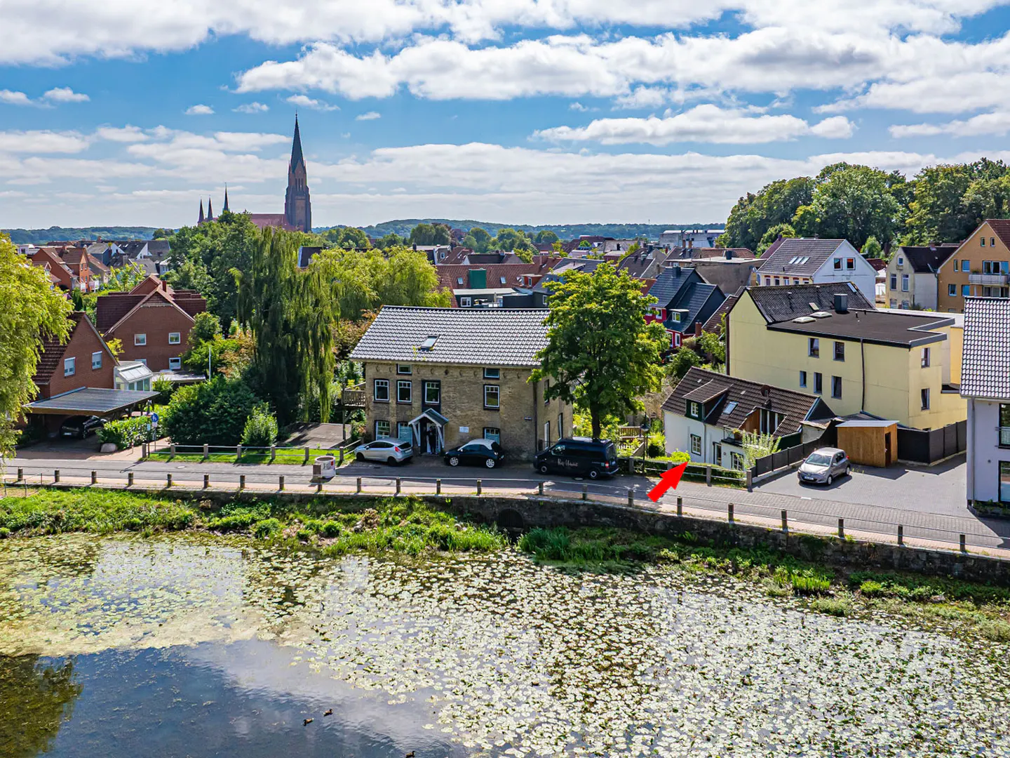 Aerial view of a European town with a church steeple, houses, cars, and a pond with lily pads. An arrow points to a white house.