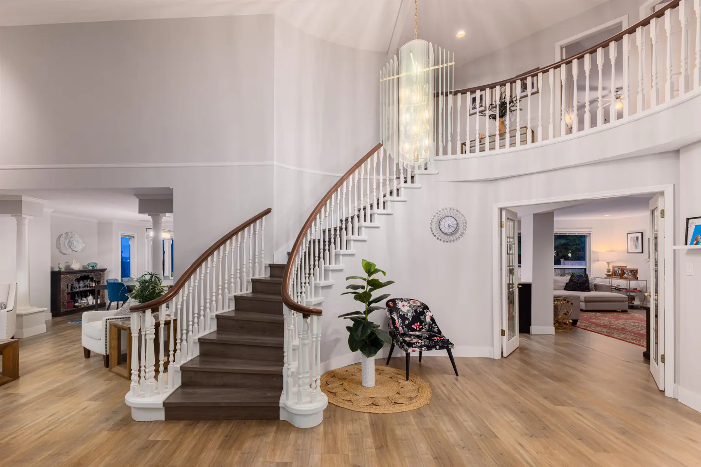 Bright foyer with wood floors, white walls, and a curved staircase with white railings. A large chandelier hangs above a floral chair and plant.
