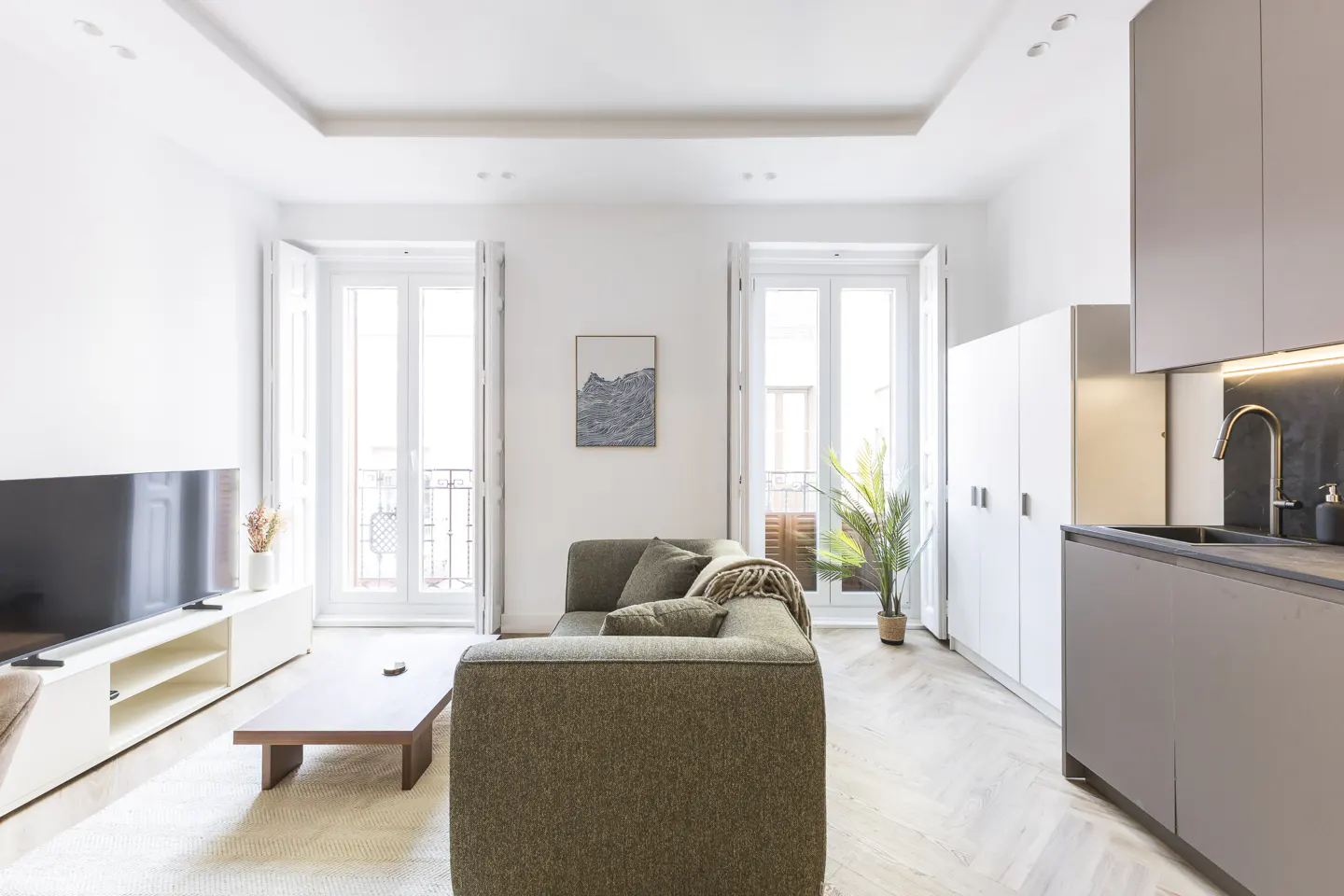Bright living room with olive sofa, wood table, TV, and kitchen area with gray cabinets and gold faucet. White walls and herringbone floors.