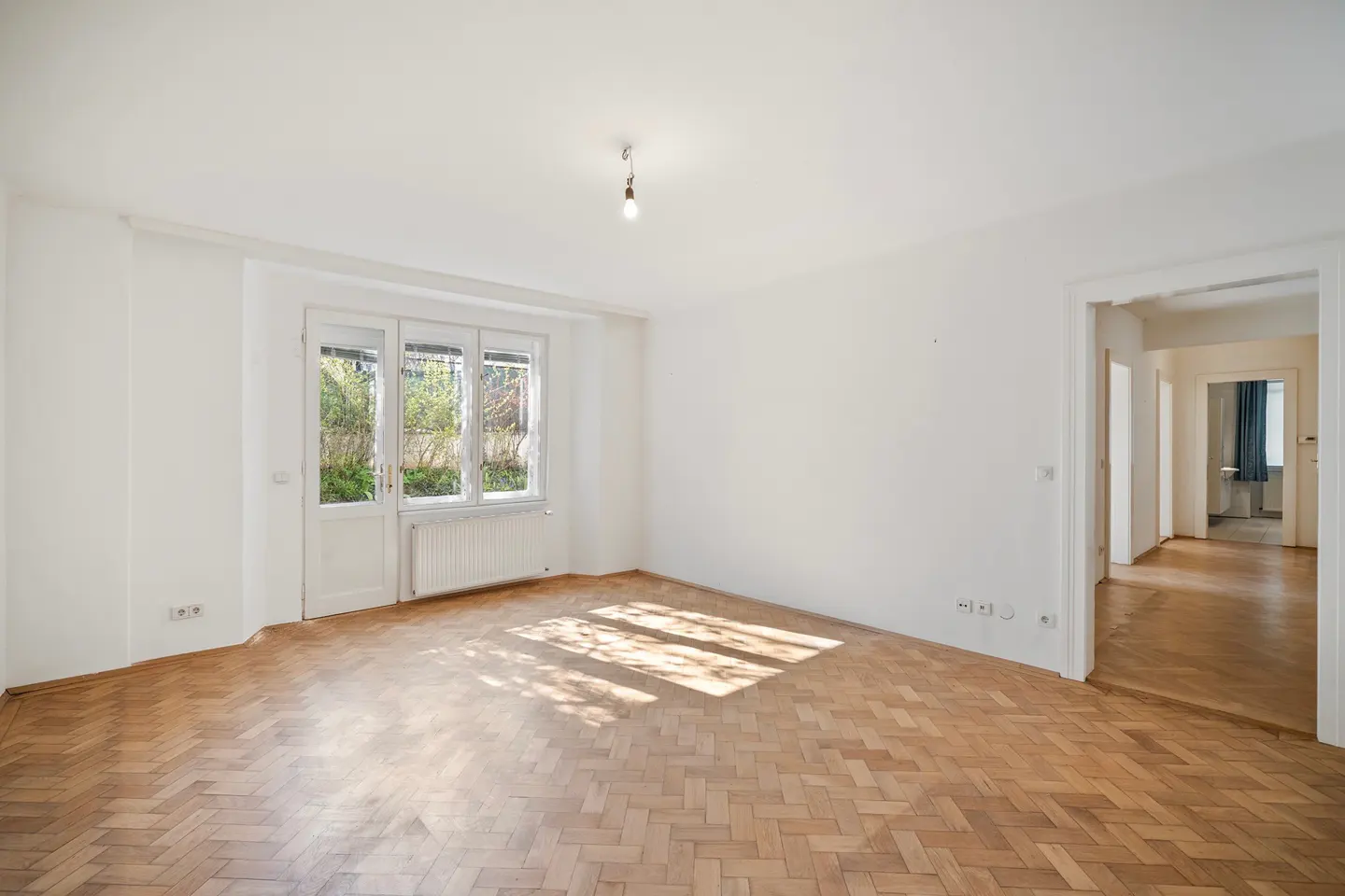 Bright, empty room with herringbone wood floors, white walls, and a window with a view of greenery. A doorway leads to another room.