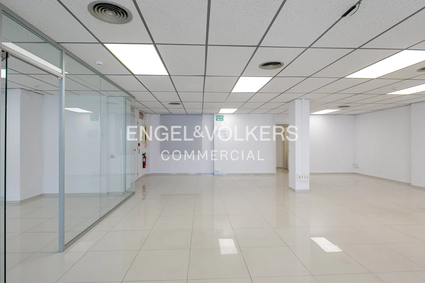 Bright, empty commercial space with white walls, tiled floor, and drop ceiling. Glass-walled office to the left. Engel & Volkers logo on wall.