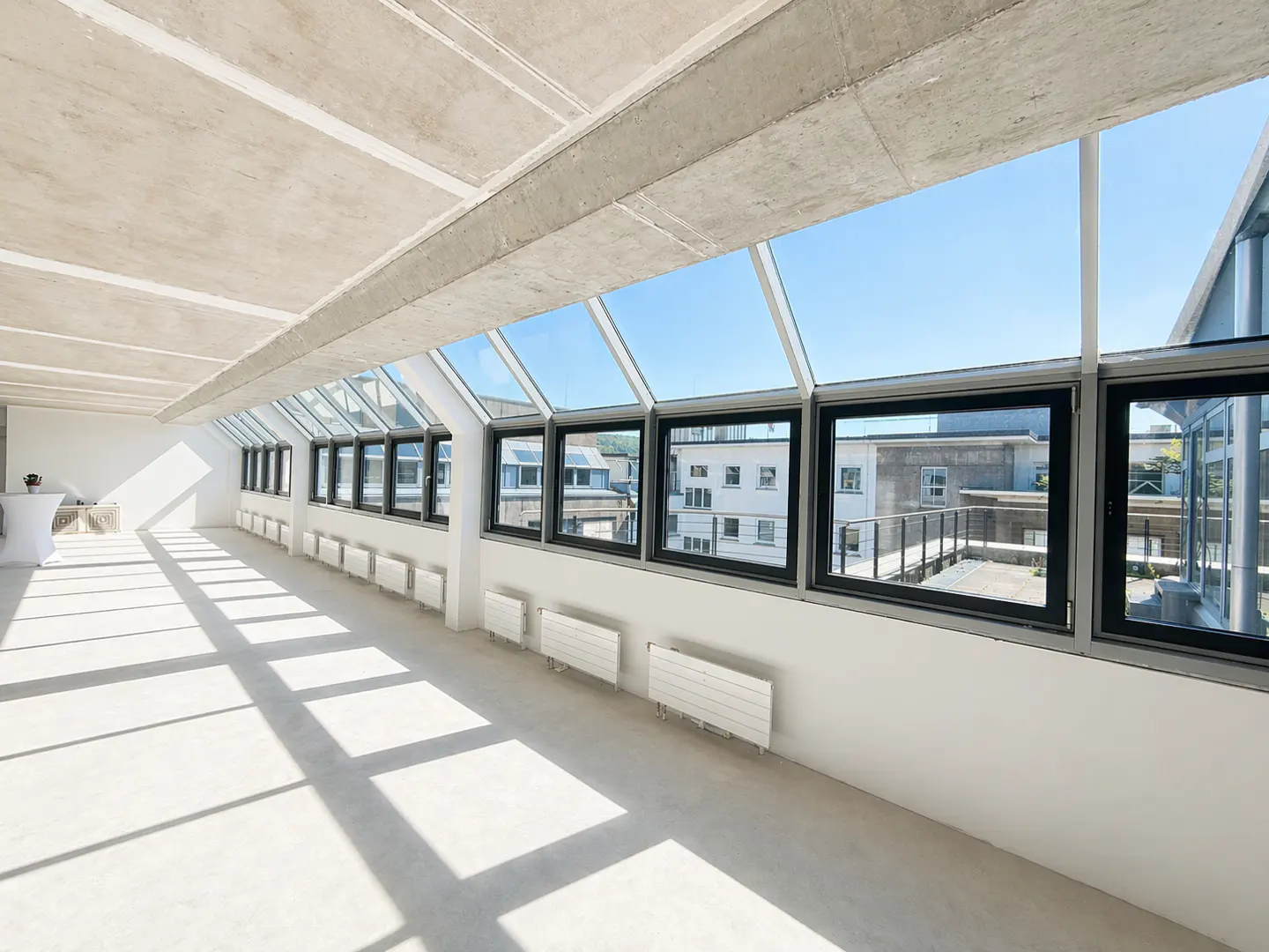 Bright, empty room with concrete ceiling and floor. Large windows with black frames line the wall, offering a view of buildings and blue sky.