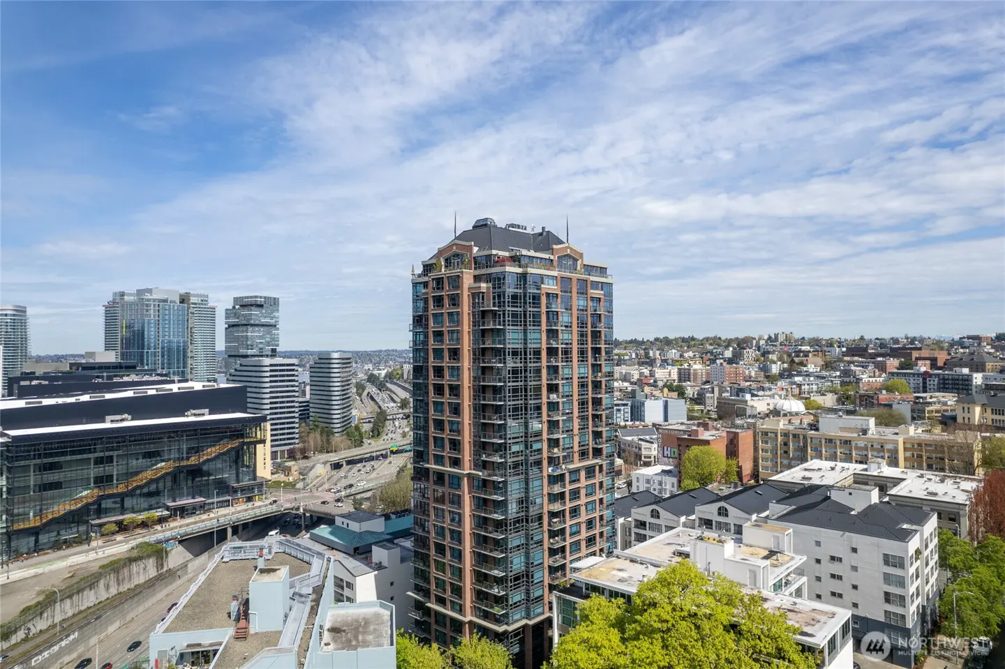 A tall, modern apartment building with glass windows and brick accents, surrounded by a cityscape under a blue sky.