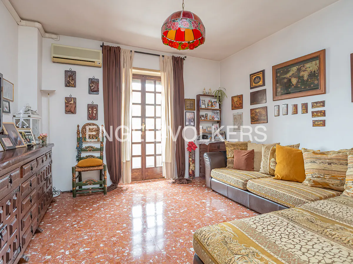 Bright living room with a brown L-shaped sofa, wooden furniture, and a red-orange patterned floor. A colorful lamp hangs from the ceiling.