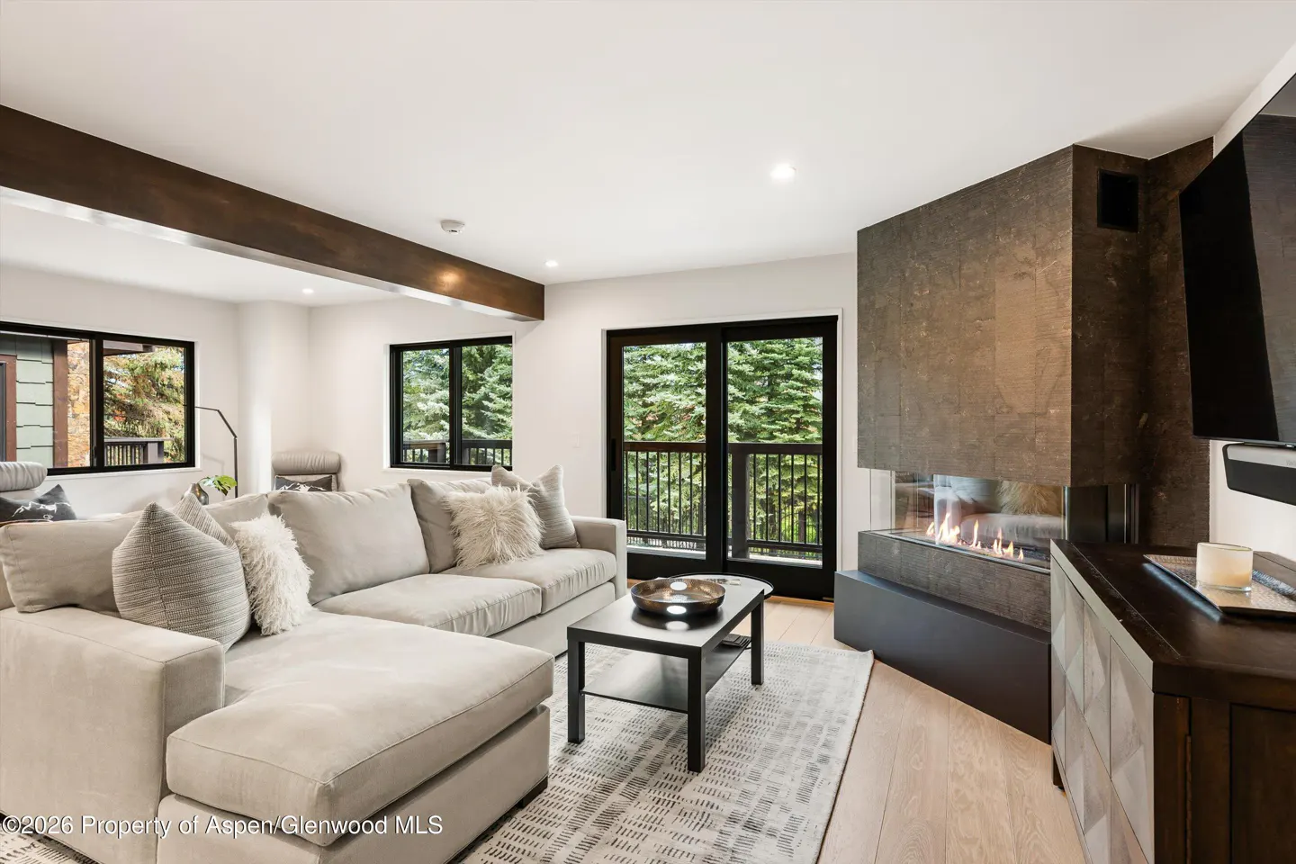 Living room with a gray sectional sofa, black coffee table, fireplace, and windows with a view of trees.