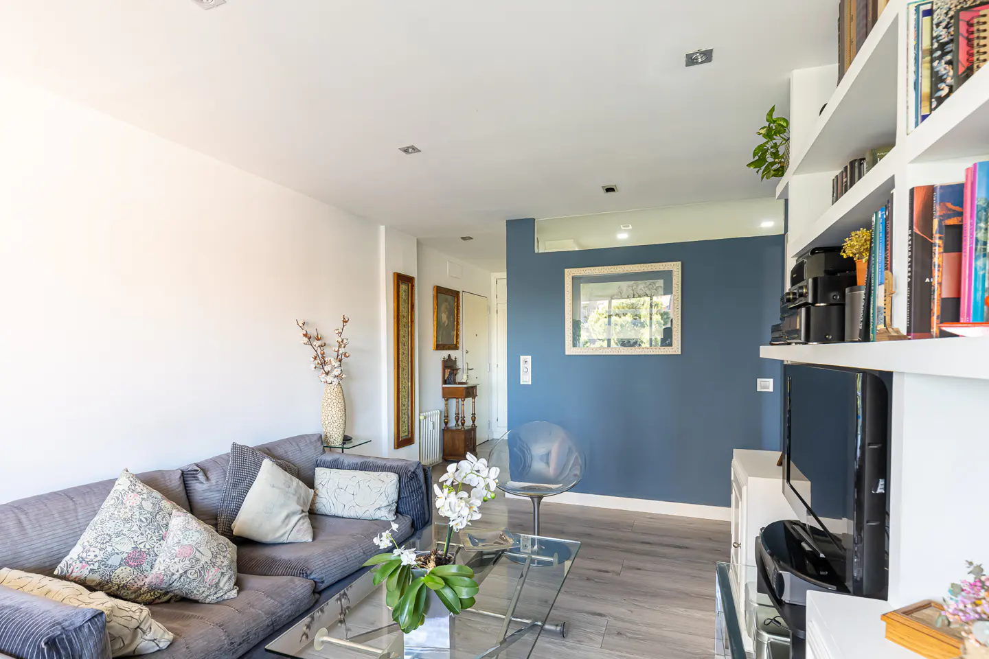 Living room with gray sofa, glass table, and white shelves filled with books and electronics. Blue accent wall with framed art.