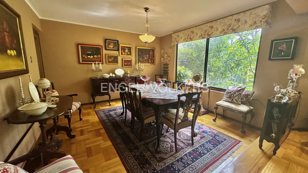 A dining room with a dark wood table and chairs on a patterned rug. A large window looks out onto green trees. Paintings adorn the tan walls.