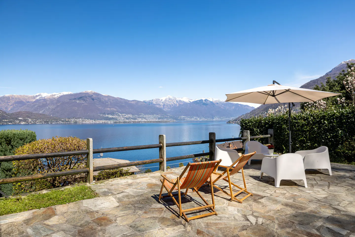 Outdoor patio with lake view. Two striped lounge chairs face the water. White chairs and umbrella sit on a stone patio. Mountains in the background.