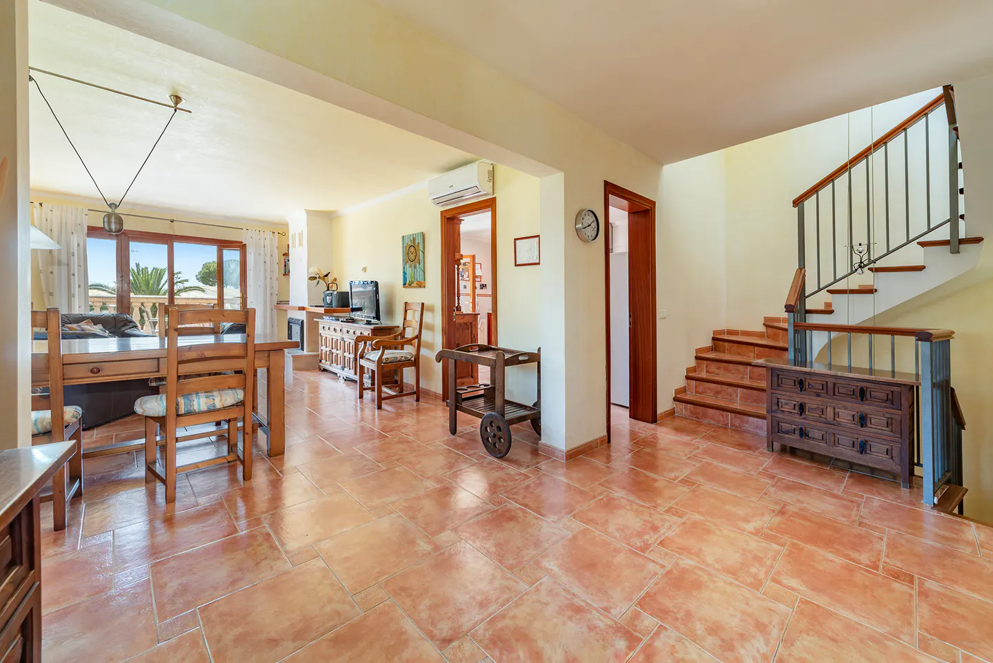 Open-concept living space with terracotta tile flooring, wooden dining set, staircase, and serving cart.