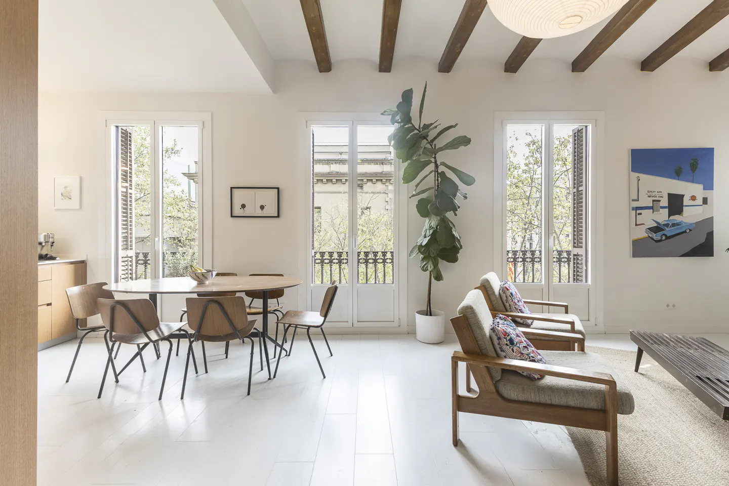 Bright, modern living space with white floors, exposed beams, and large windows. A dining table, chairs, and armchairs furnish the room.