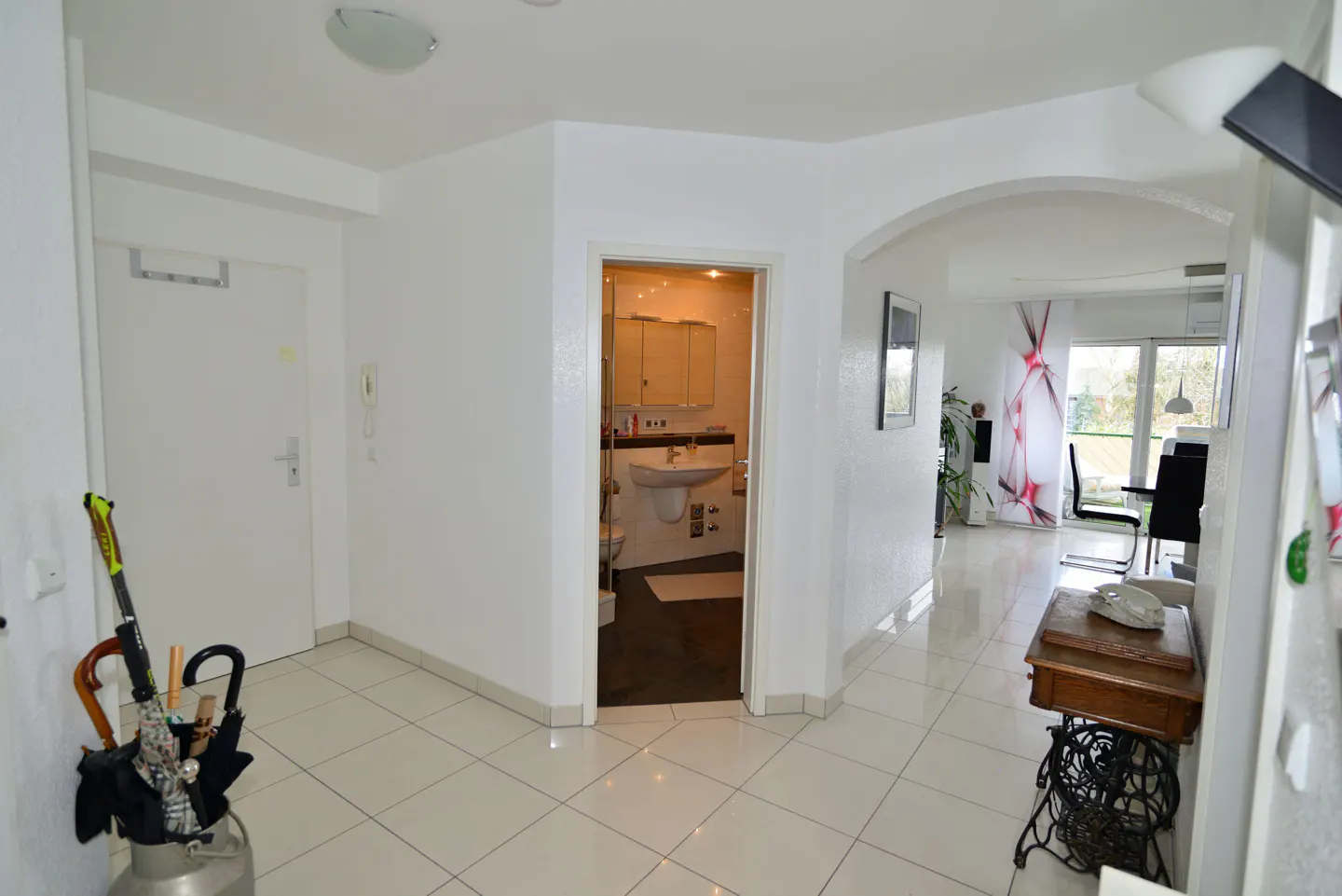 Apartment entryway with white tile floors, white walls, and open doorways to a bathroom and living area. An umbrella stand sits by the front door.