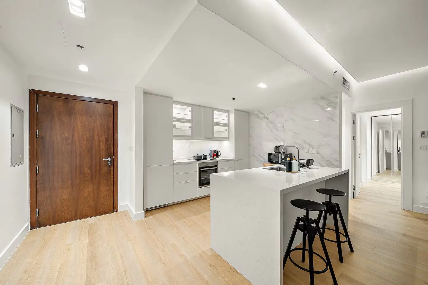 Bright, modern kitchen with white cabinets, marble backsplash, and island with black stools. A wood door is on the left.