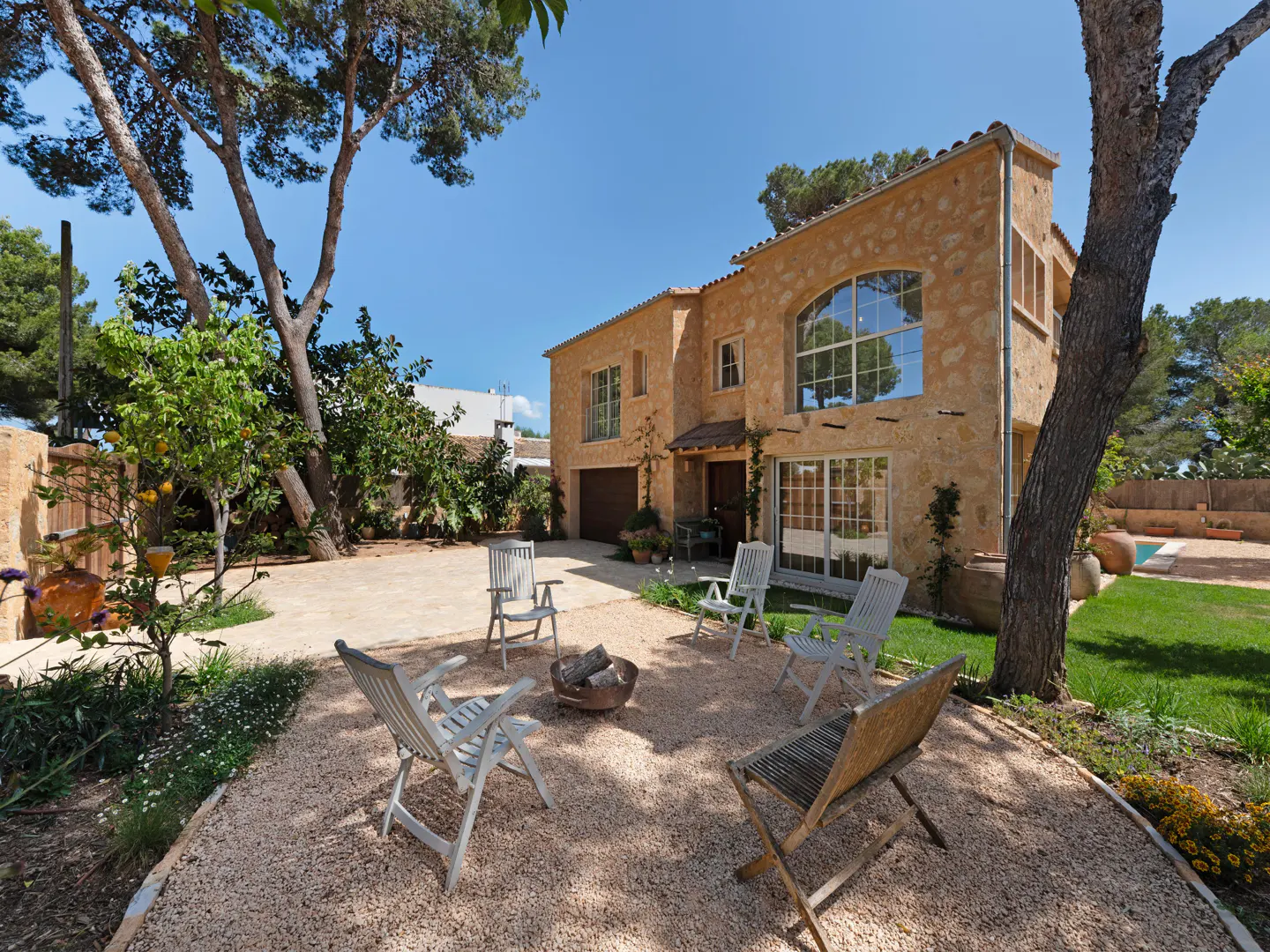 Stone house with a gravel courtyard, surrounded by trees and greenery. Chairs are arranged around a fire pit. Blue sky.