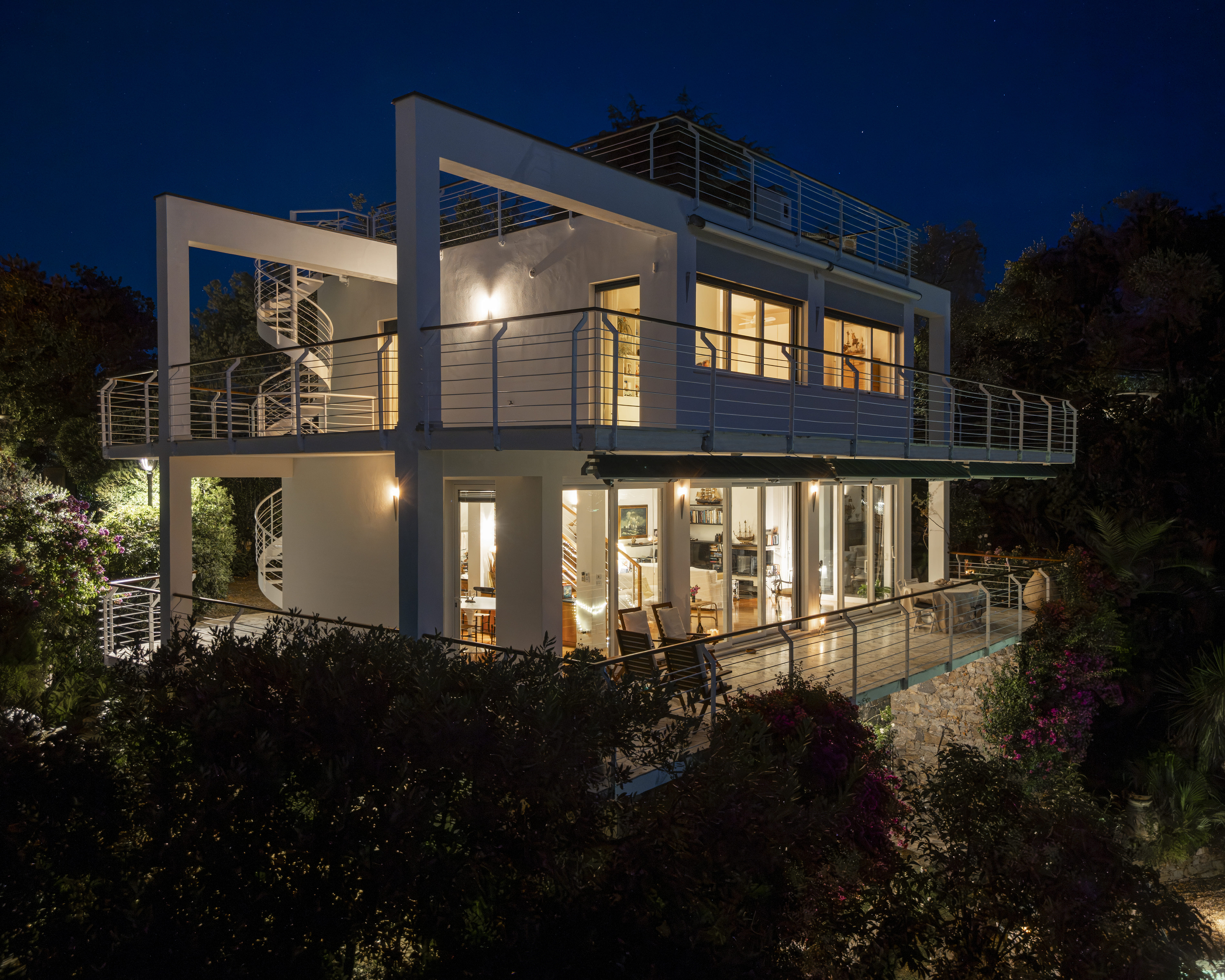 Modern white two-story house with spiral staircases and illuminated balconies at night, surrounded by lush landscaping. [AI-generated text]