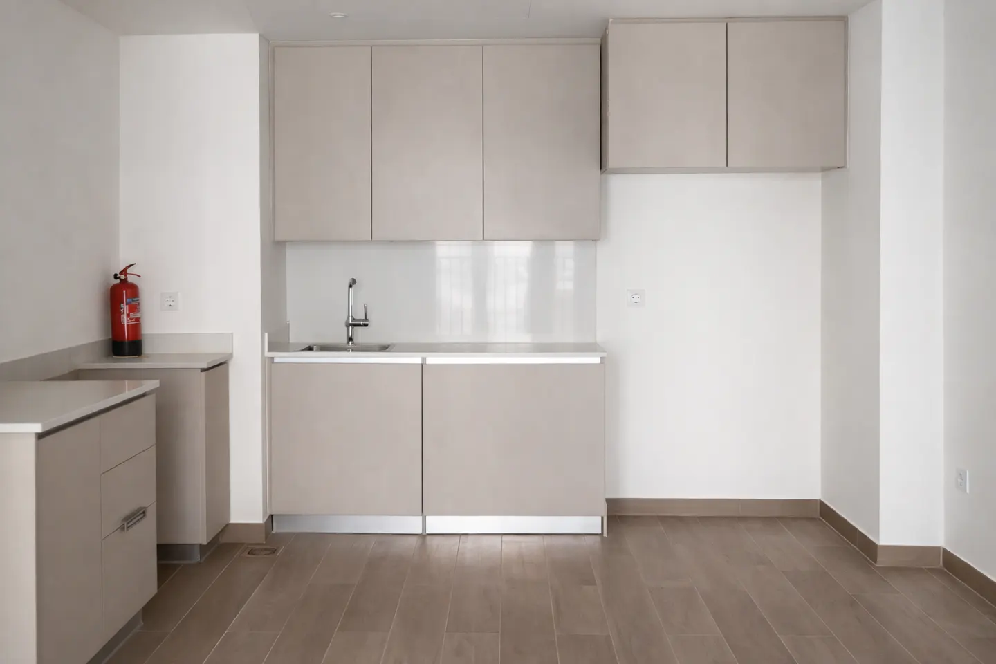 A bright, empty kitchen with light brown cabinets, a stainless steel sink, and wood floors. A red fire extinguisher is visible.