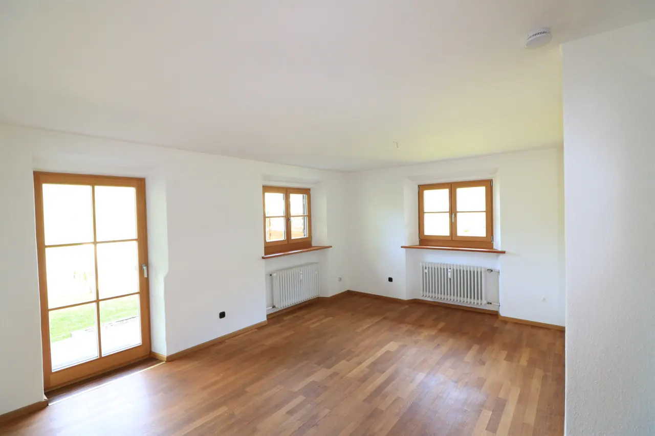 Bright, empty room with hardwood floors, white walls, and wood-framed windows. Radiators sit beneath the windows. A glass door lets in natural light.