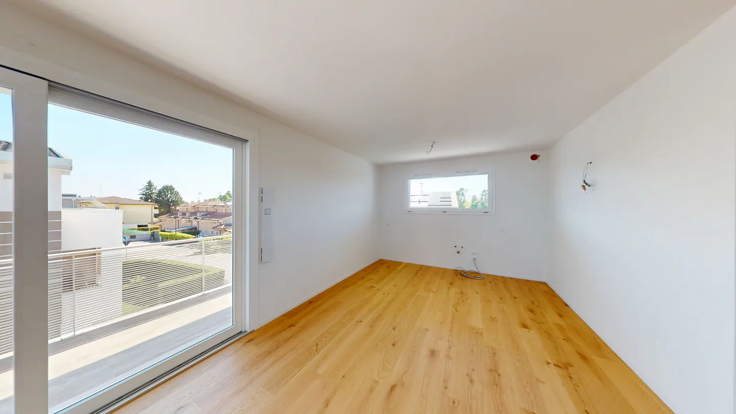 Bright, empty room with light wood floors and white walls. Large sliding glass door leads to a balcony with a view of houses.