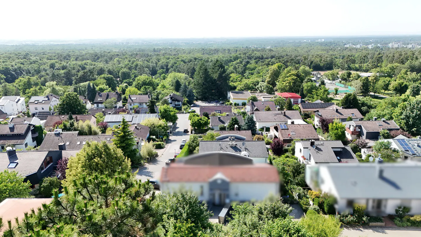 Aerial view of a suburban neighborhood with houses, trees, and a forest in the background. Some houses have solar panels.