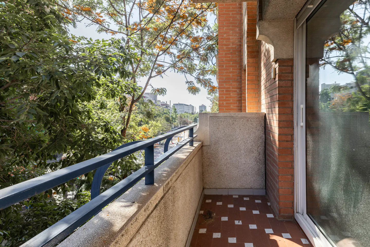 Balcony view with blue railing, brick walls, and patterned tile floor. Trees and city buildings visible in the background.