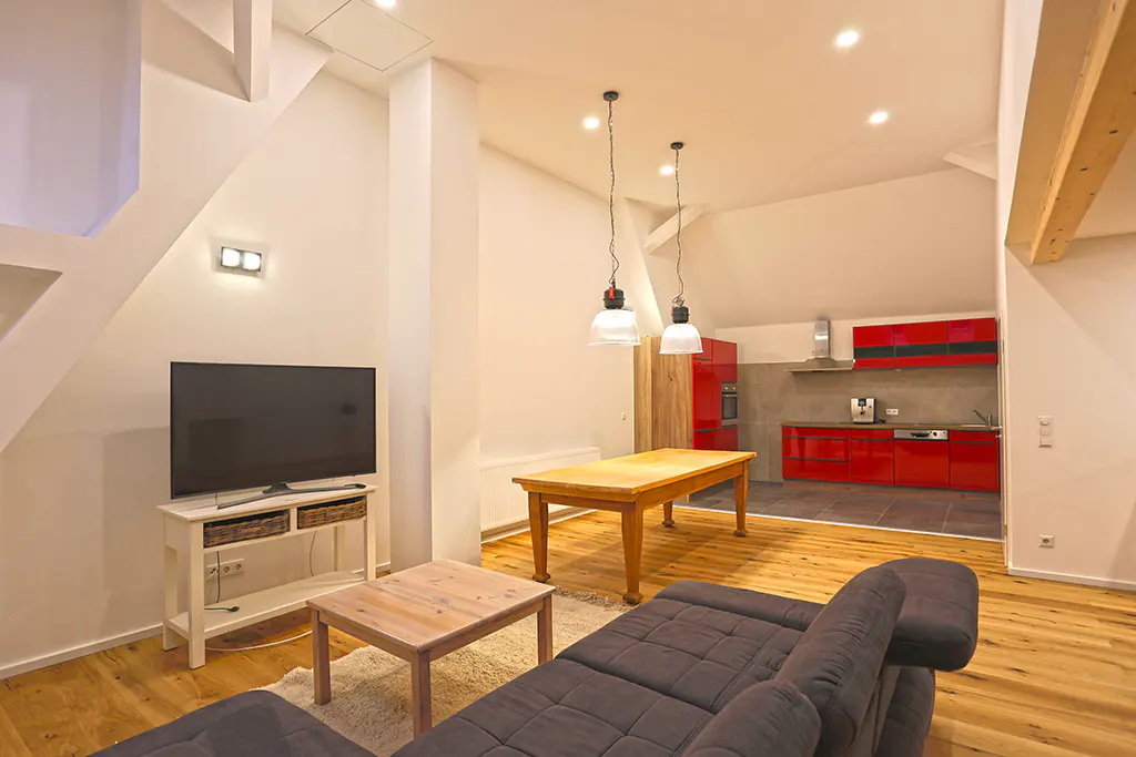 Living room with a gray sofa, wooden tables, and a TV. A red kitchen is visible in the background.