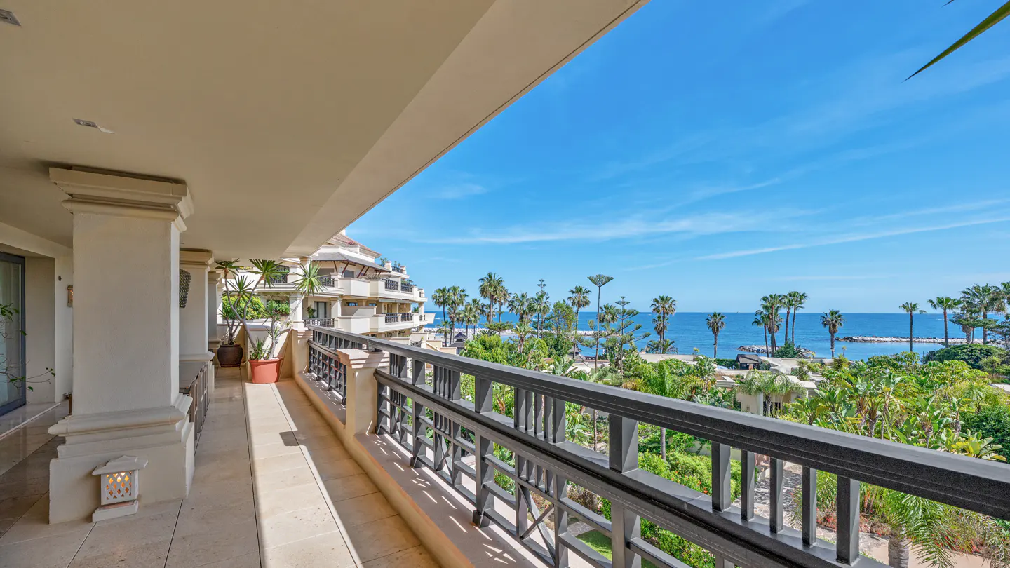 Balcony view with black railings overlooking palm trees, blue ocean, and sky. Cream-colored building with pillars on the left.