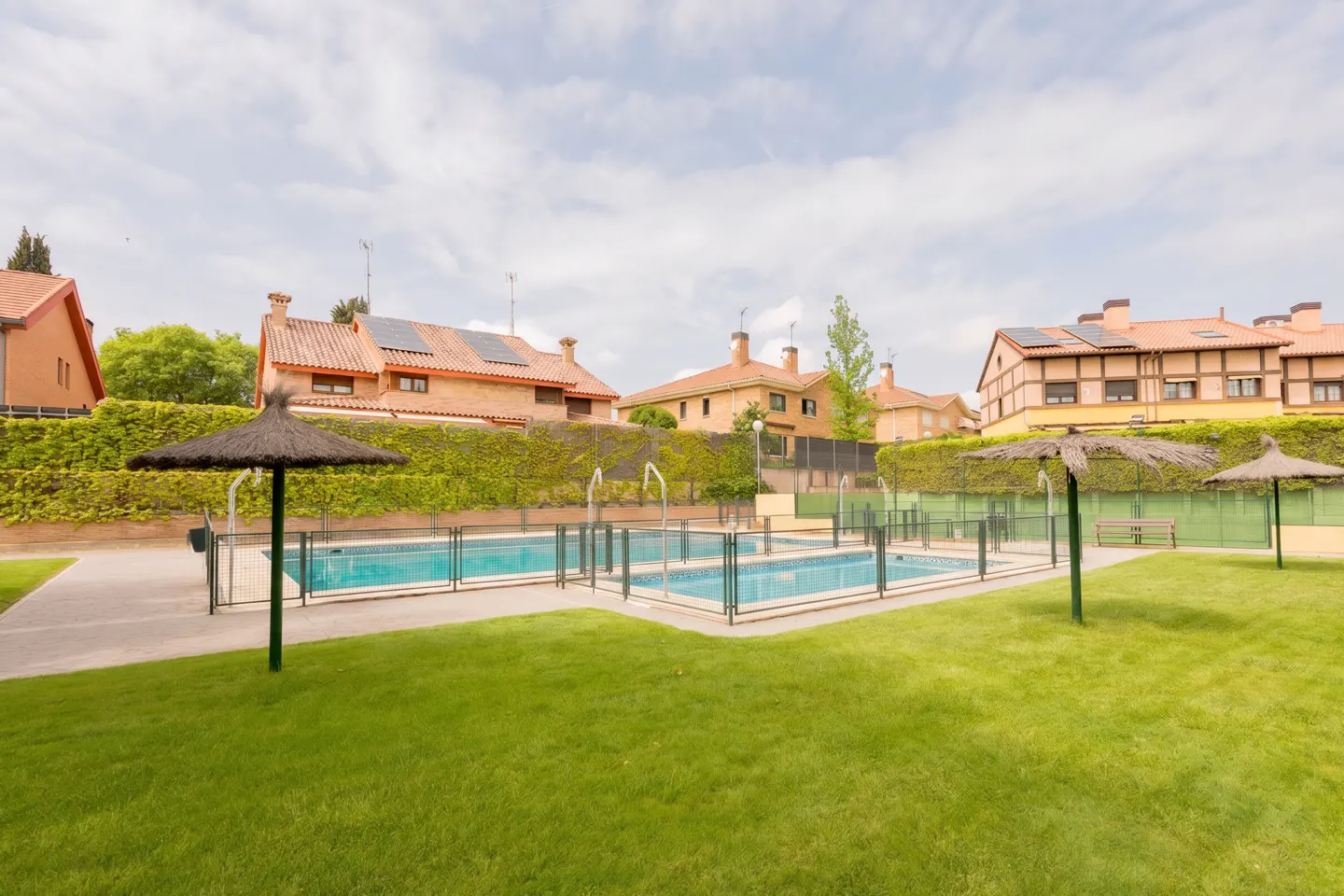Two fenced pools sit on green grass, with straw umbrellas. Brick houses with red roofs are in the background.