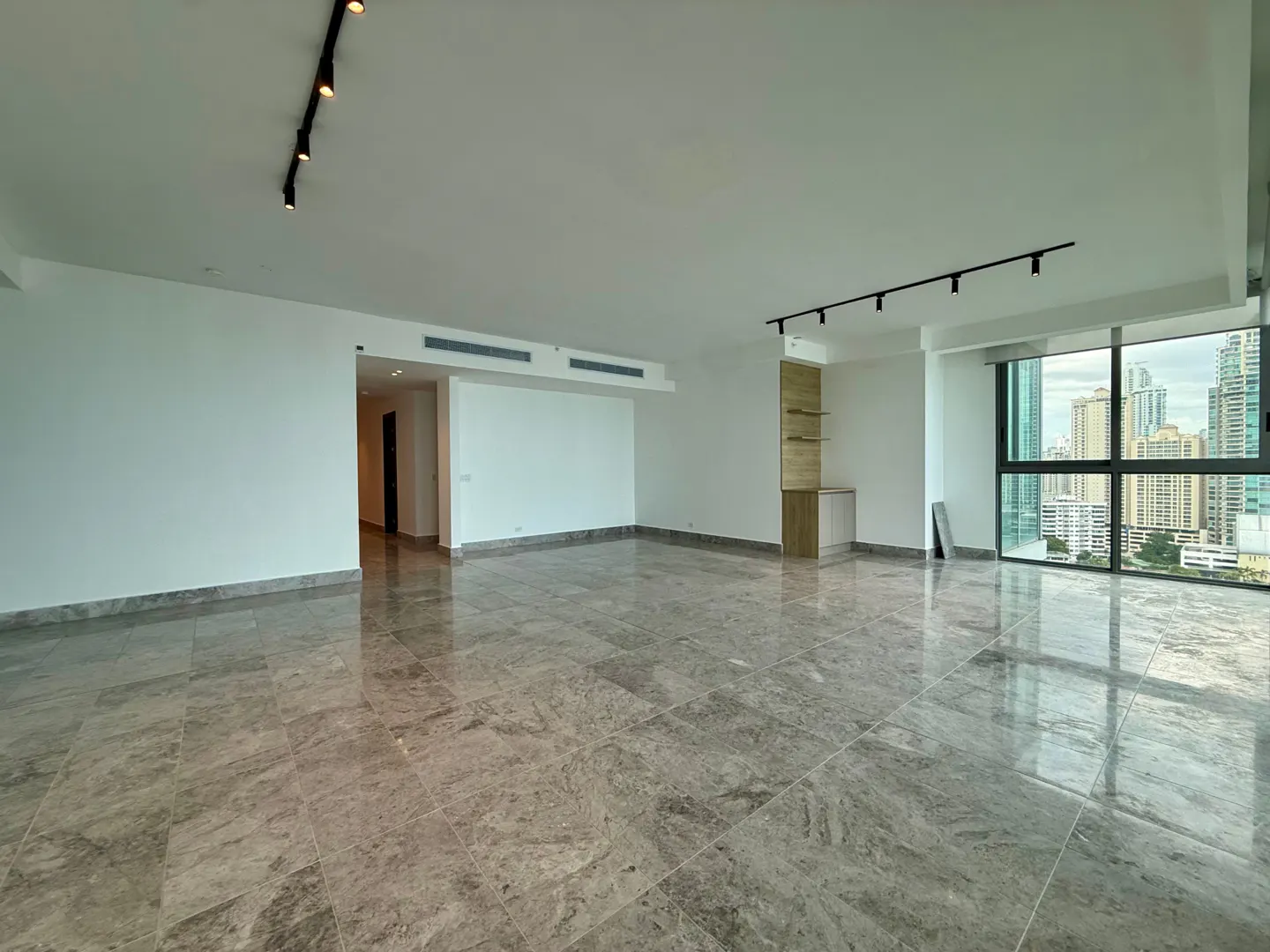 An empty, modern apartment with gray marble floors, white walls, track lighting, and a city view from a large window.