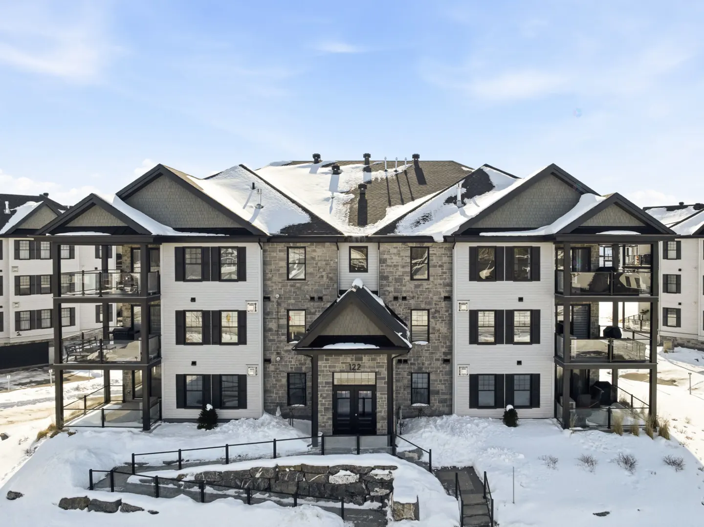 Three-story apartment building with snow-covered roof and ground. White siding, black shutters, and stone accents. Balconies with glass railings.