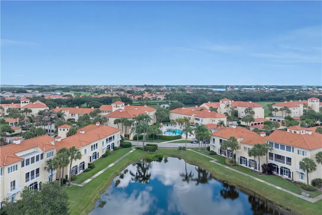 Aerial view of a residential community with beige buildings, red tile roofs, palm trees, a pool, and a pond reflecting the blue sky.
