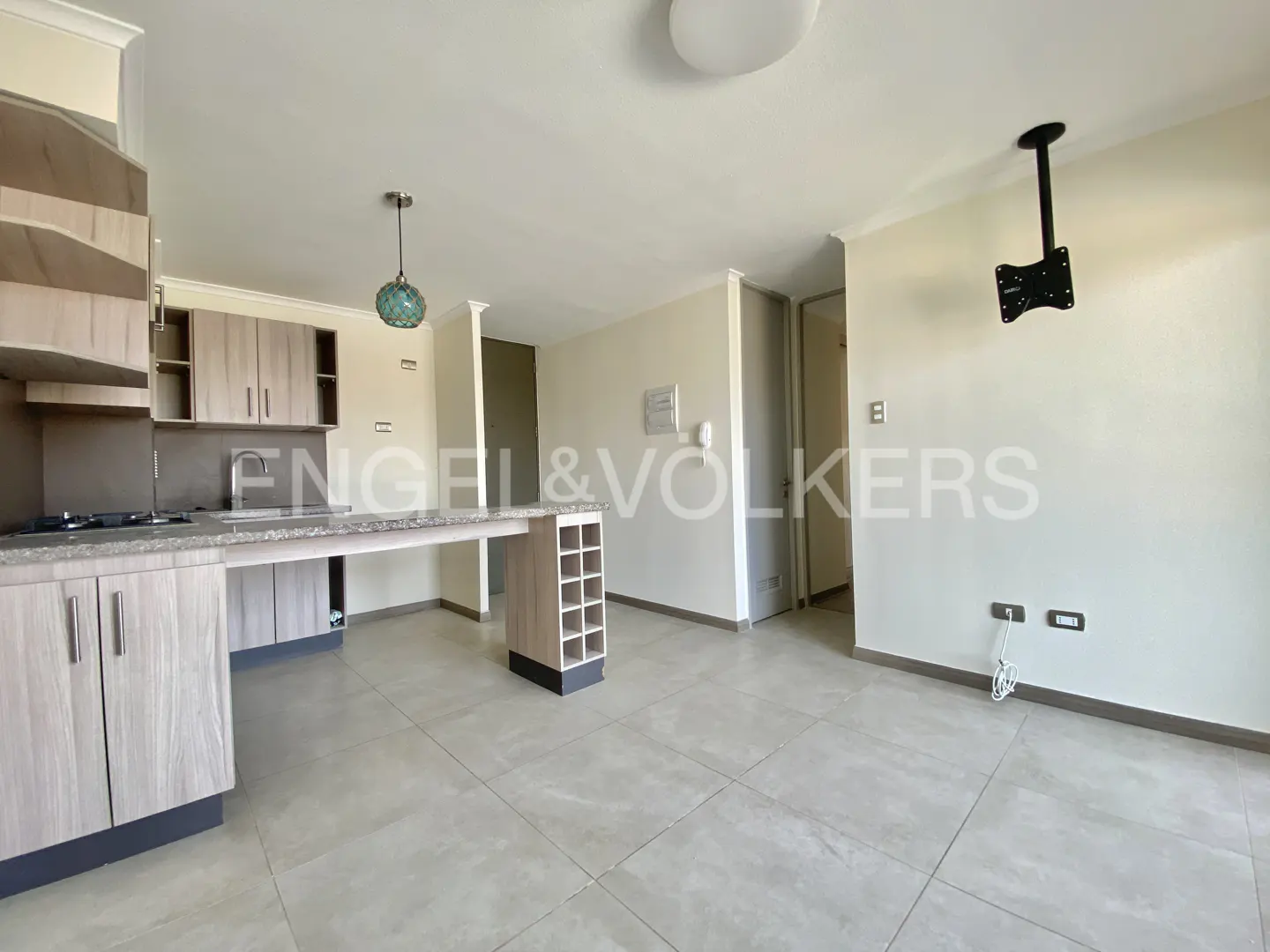 A bright, open-concept kitchen and living area with light wood cabinets and gray tile flooring. A TV mount hangs from the ceiling.