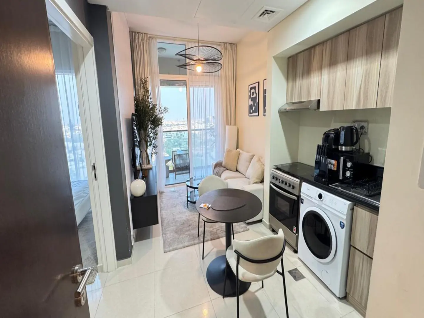 A modern apartment interior with a living area, kitchen, and balcony. Neutral tones, a round table, and city views are visible.
