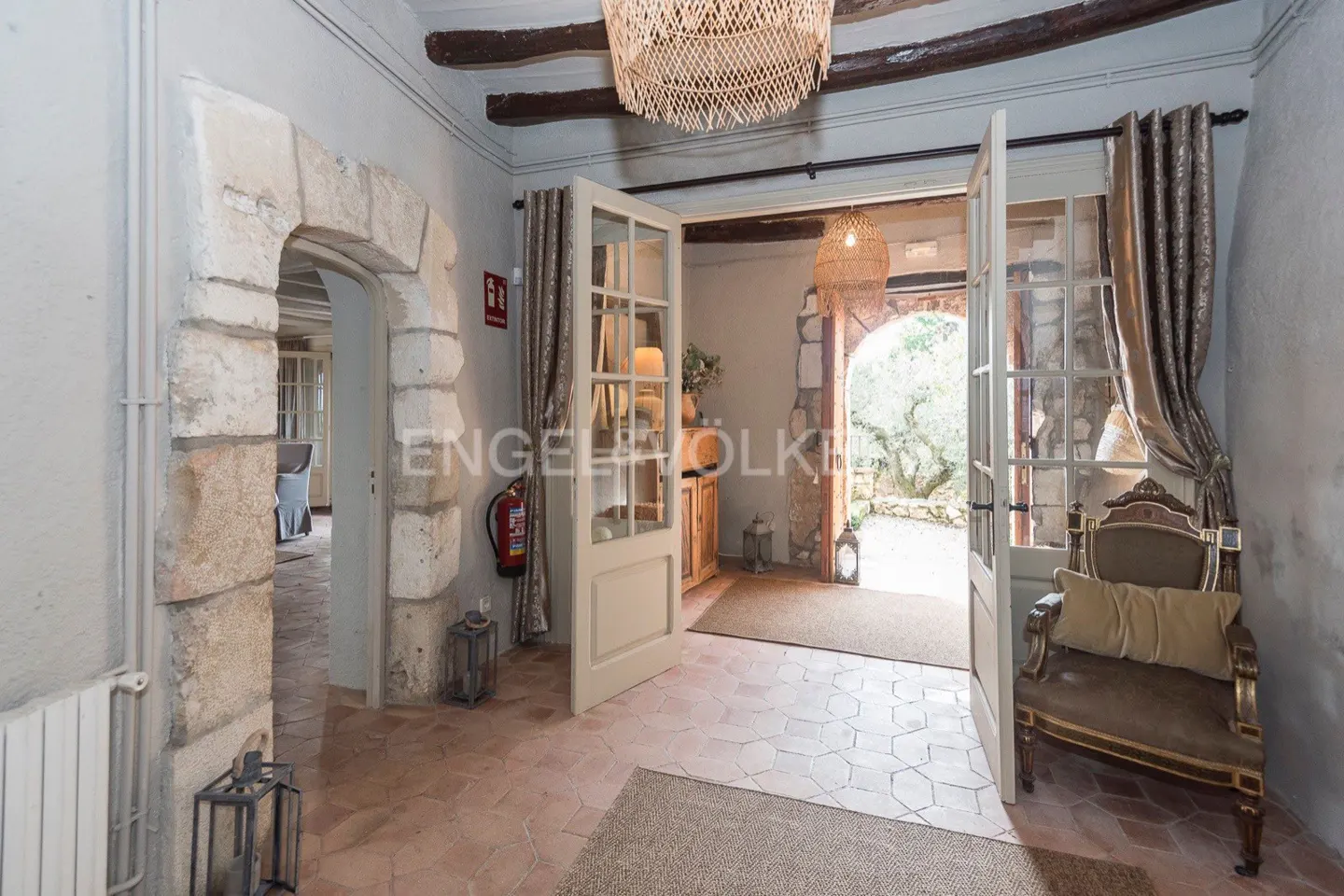 Interior view of a home's entrance with stone archways, open white doors, and a decorative chair. A woven chandelier hangs from the ceiling.