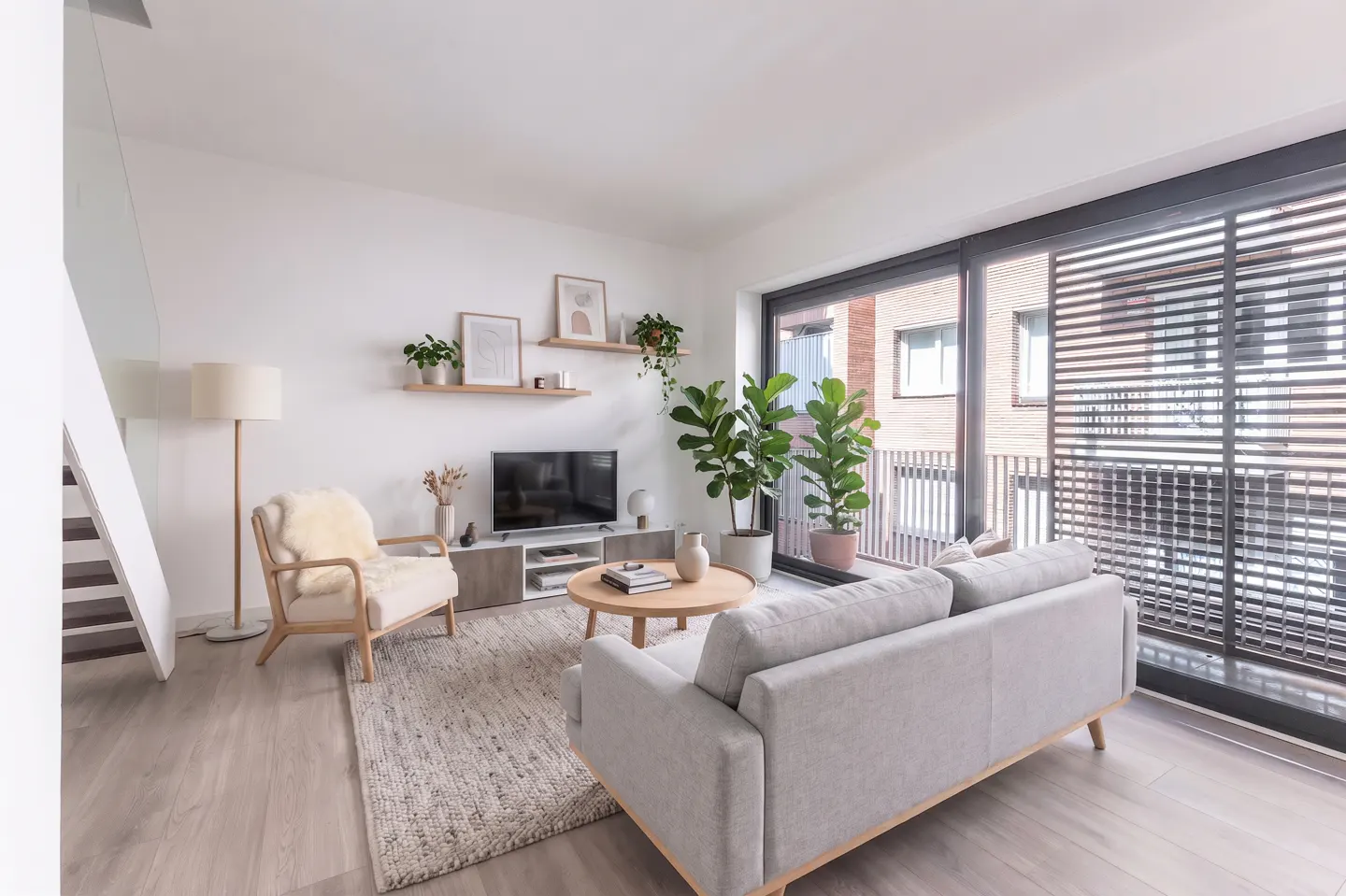 Bright living room with a gray sofa, wood chair, TV, and plants. Large sliding glass doors lead to a balcony.