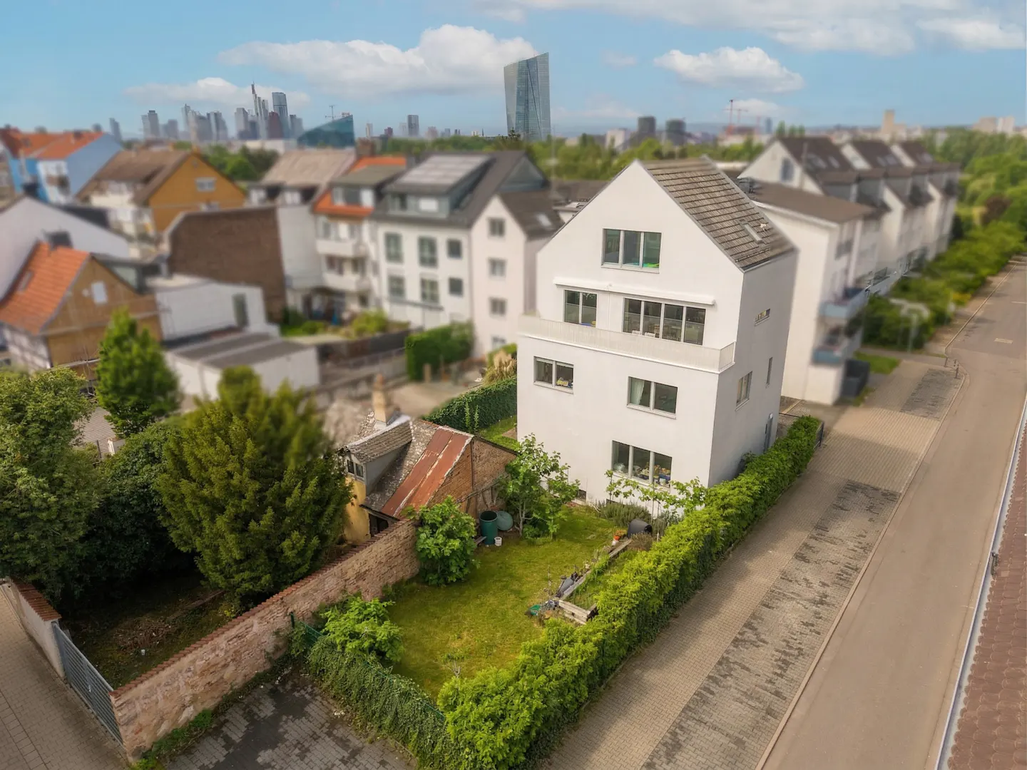 Aerial view of a modern, three-story white house with a green lawn and hedge, with a city skyline in the background.