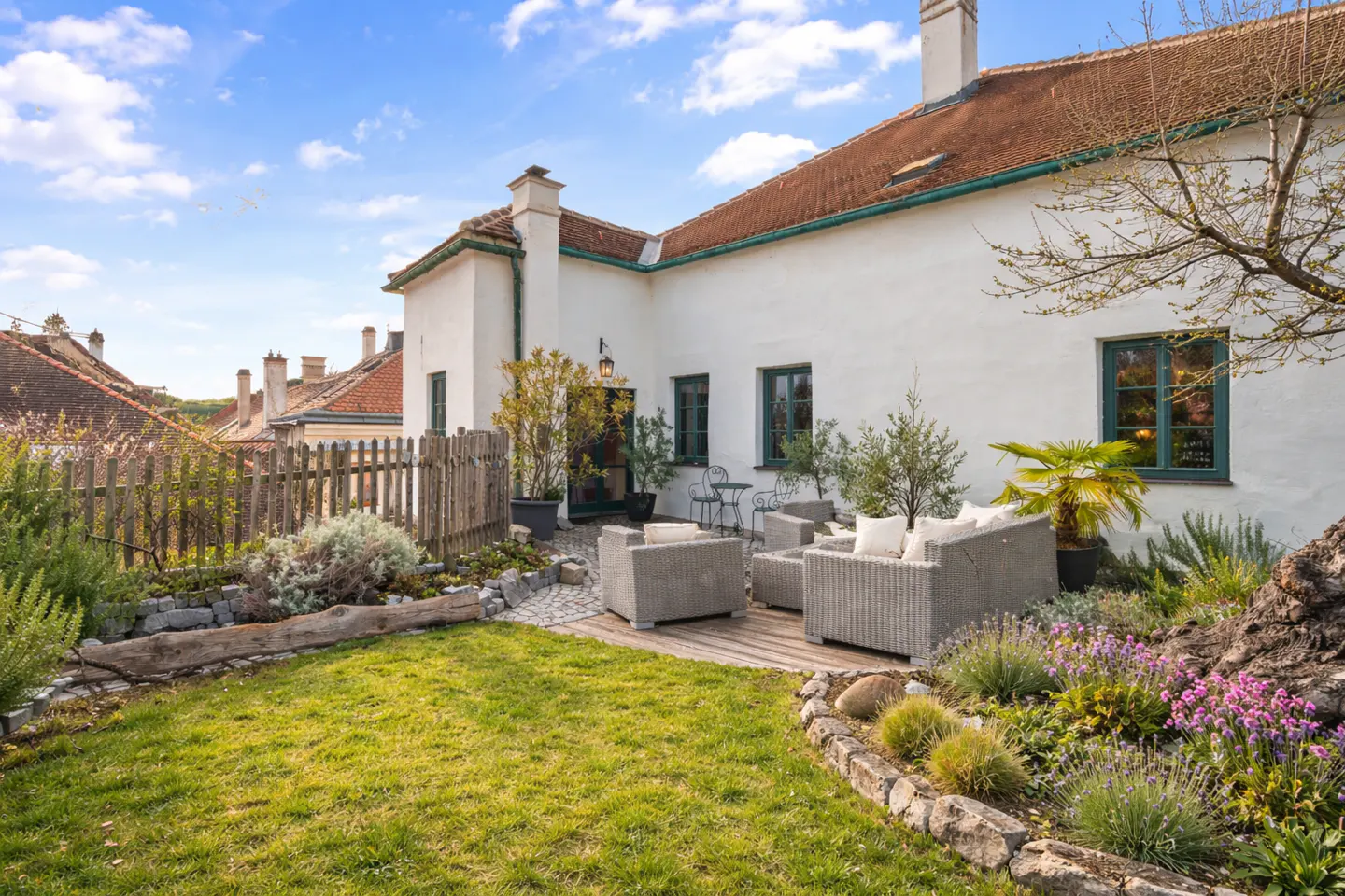 Exterior view of a white house with a red roof, green trim, and a patio with gray wicker furniture. A green lawn and flower garden are in the foreground.