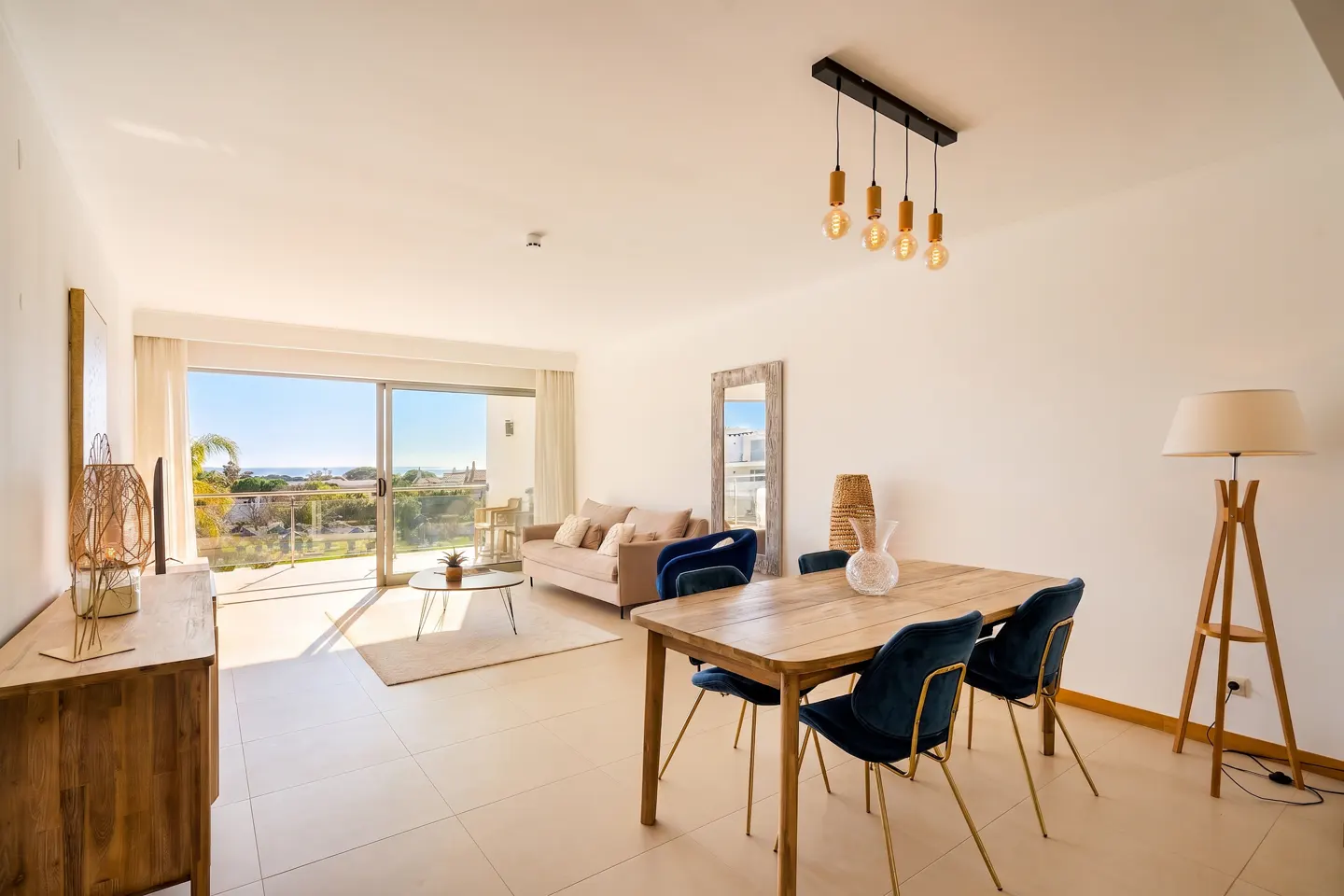 Bright, modern living room with a wood dining table, blue chairs, and a beige sofa. Sliding glass doors open to a balcony with ocean views.