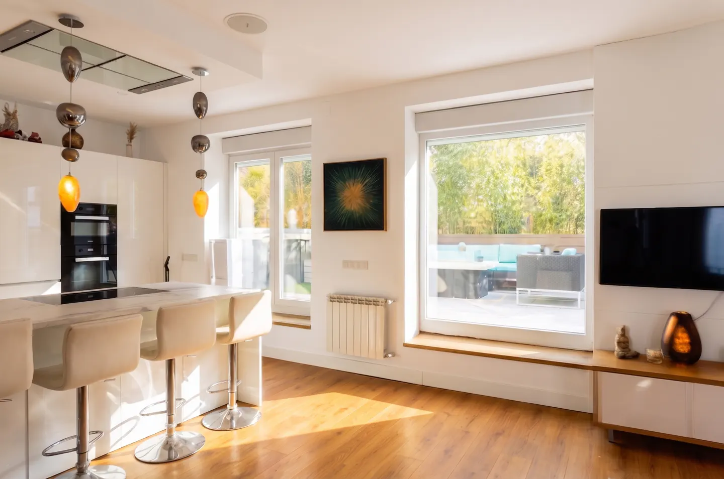 Bright, modern kitchen with white cabinets, island with stools, and large windows overlooking an outdoor patio. Hardwood floors and pendant lights.