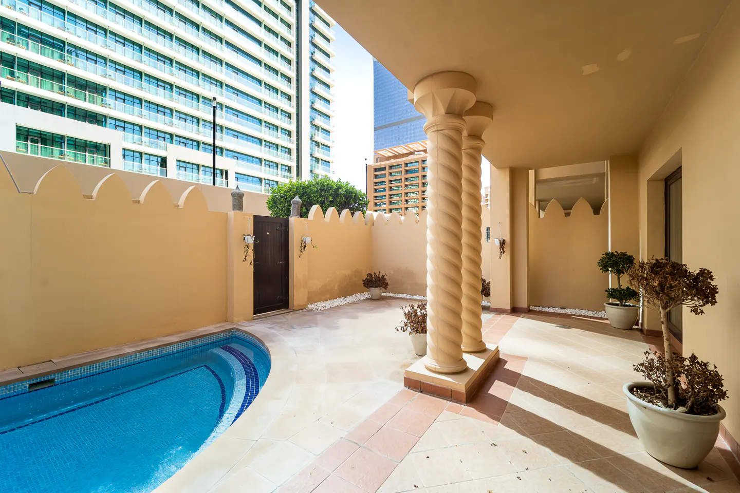 Outdoor patio with a small pool, tan walls, and spiral columns. A tall building is visible in the background.
