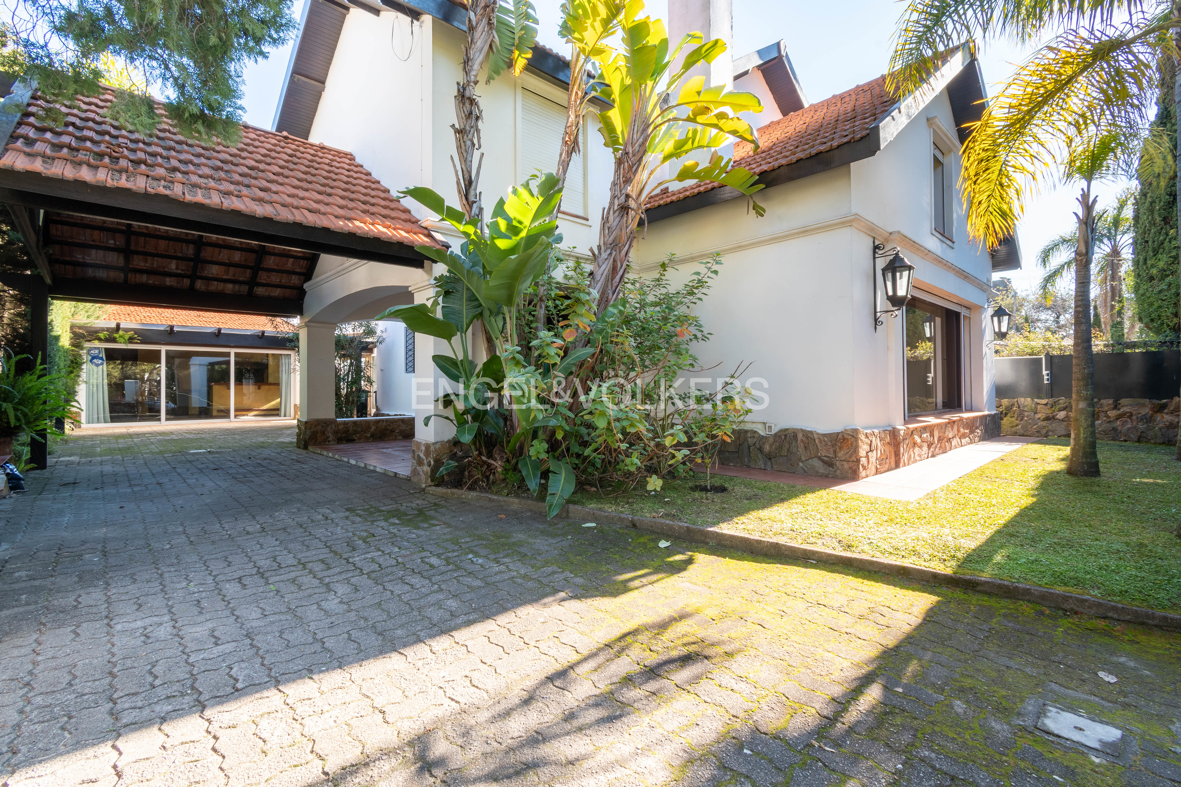 White house with a terracotta roof, paved driveway, covered carport, and lush tropical plants in a sunny garden.