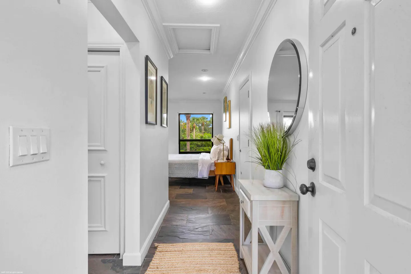 Hallway view with white walls, slate floors, and a jute rug. A round mirror hangs above a white console table with a green plant. A bedroom is visible at the end.