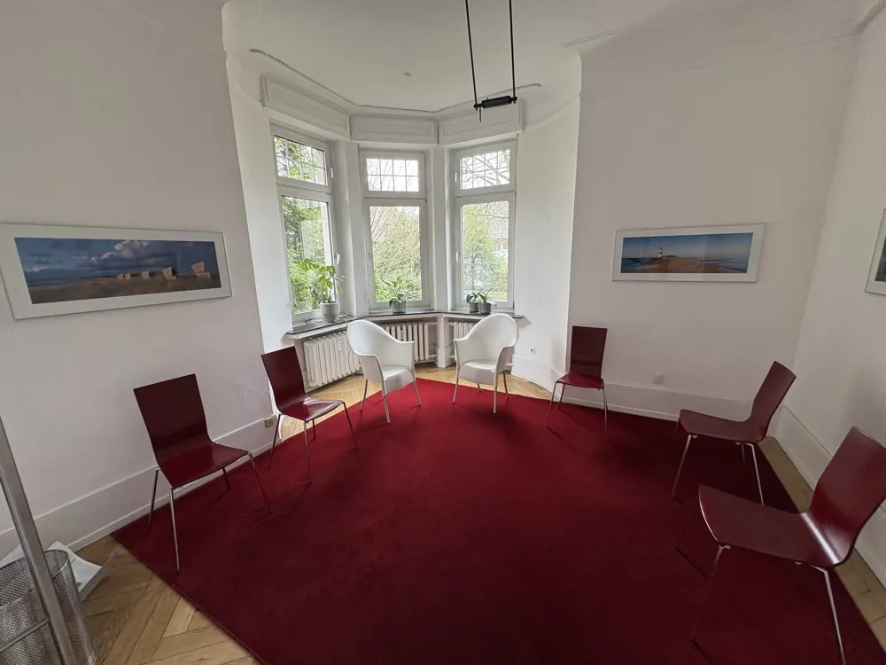 Bright room with red carpet, white walls, and wood floors. Two white chairs face a bay window with plants. Red chairs line the walls.