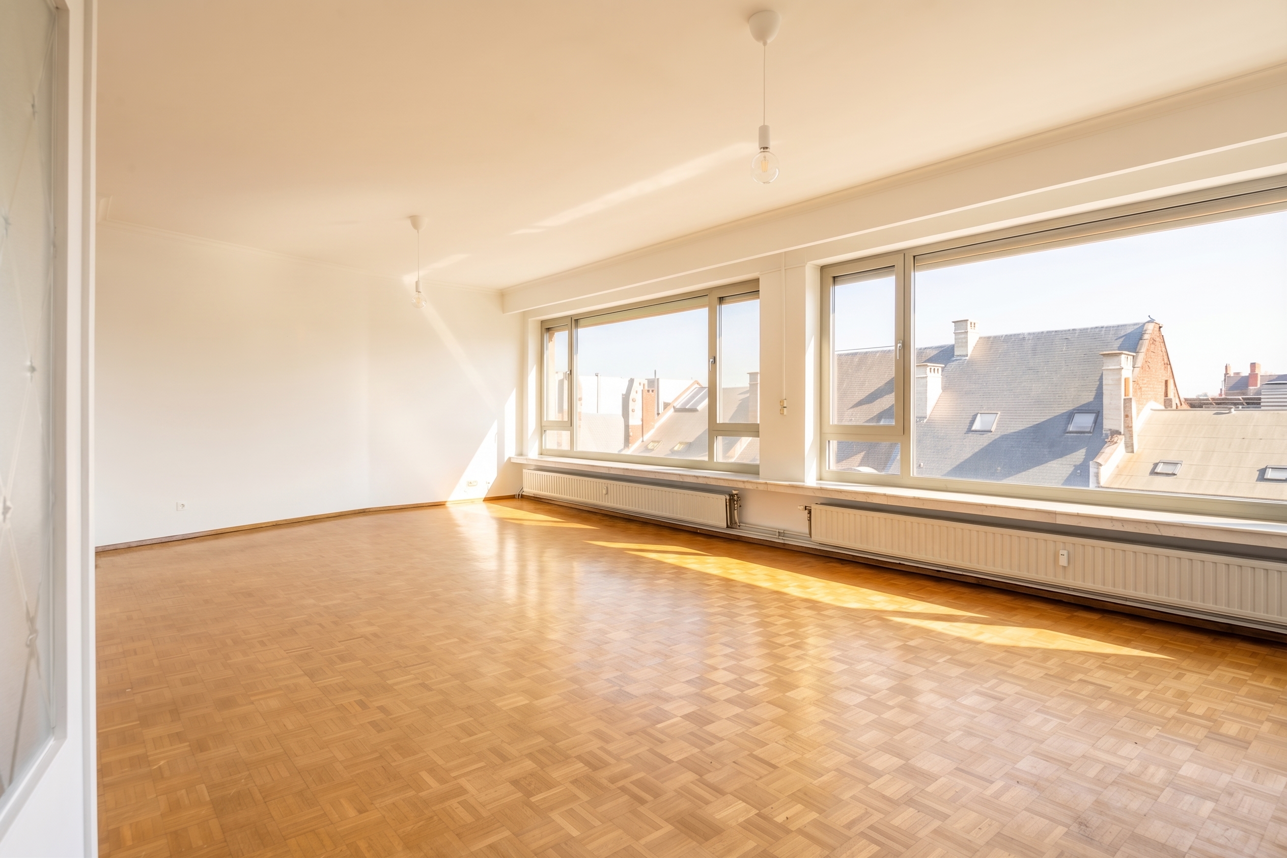 Bright, empty room with parquet floors, white walls, and large windows overlooking rooftops.
