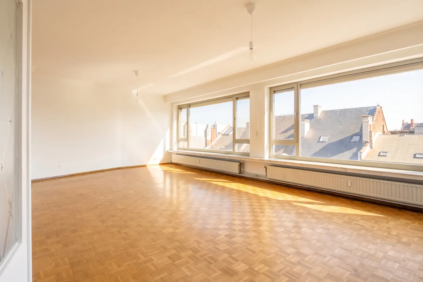 Bright, empty room with parquet floors, white walls, and large windows overlooking rooftops.