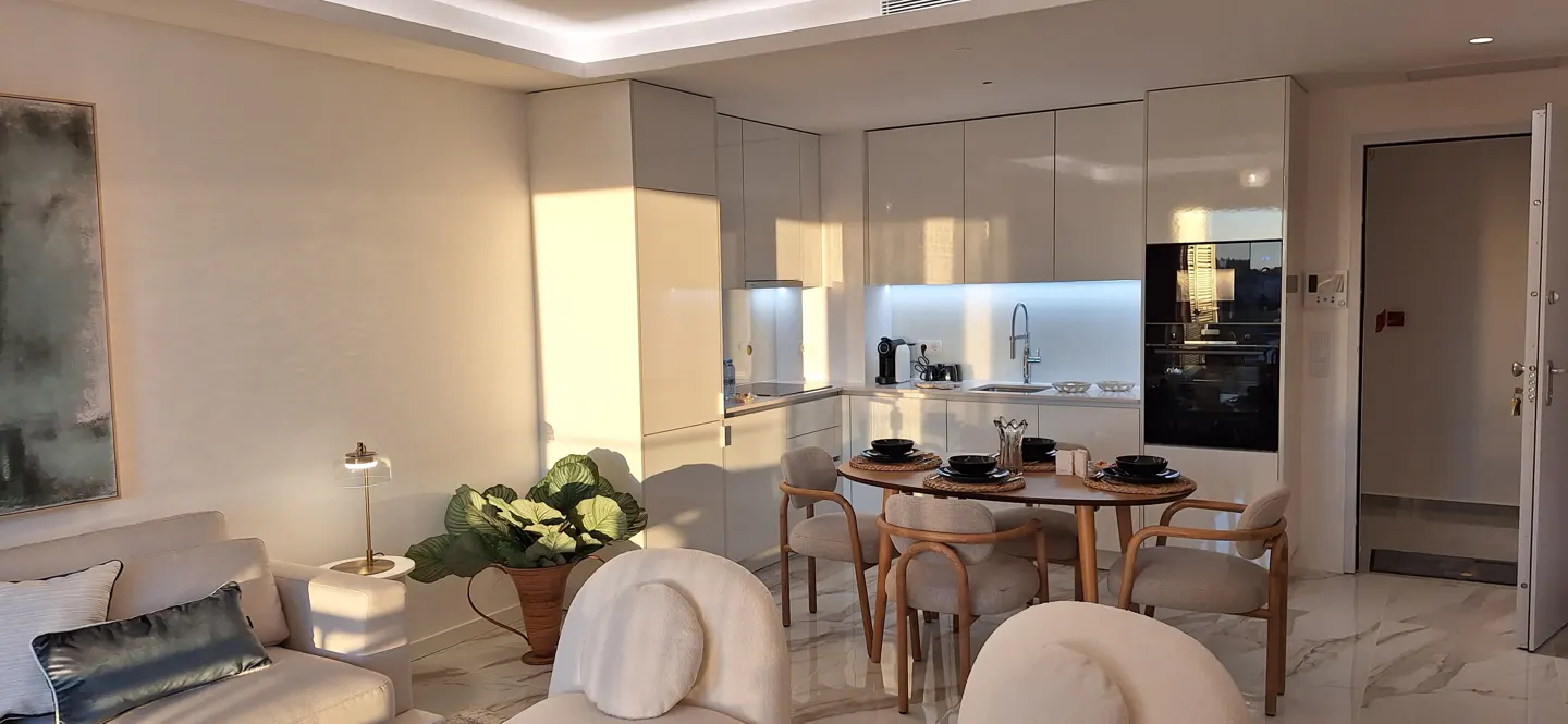 A modern apartment interior with white walls, cabinets, and marble floors. A round dining table is set for a meal, with a sofa and plant in the foreground.