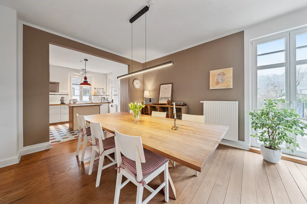 A dining room with a long wooden table, white chairs with red cushions, and a modern light fixture. A kitchen is visible through an opening.