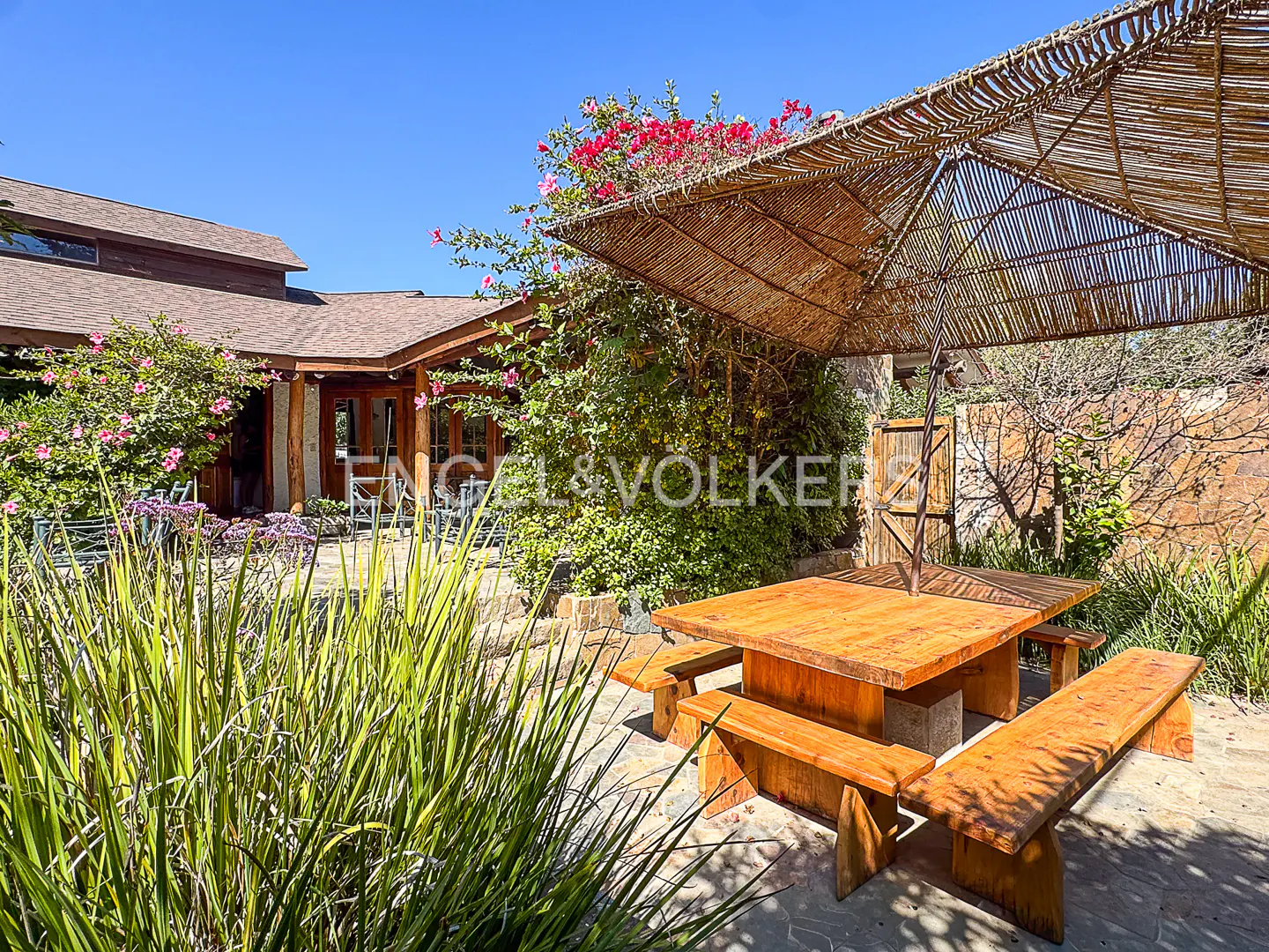 Outdoor patio with a wooden picnic table and benches under a thatched umbrella, surrounded by lush greenery and a house in the background.