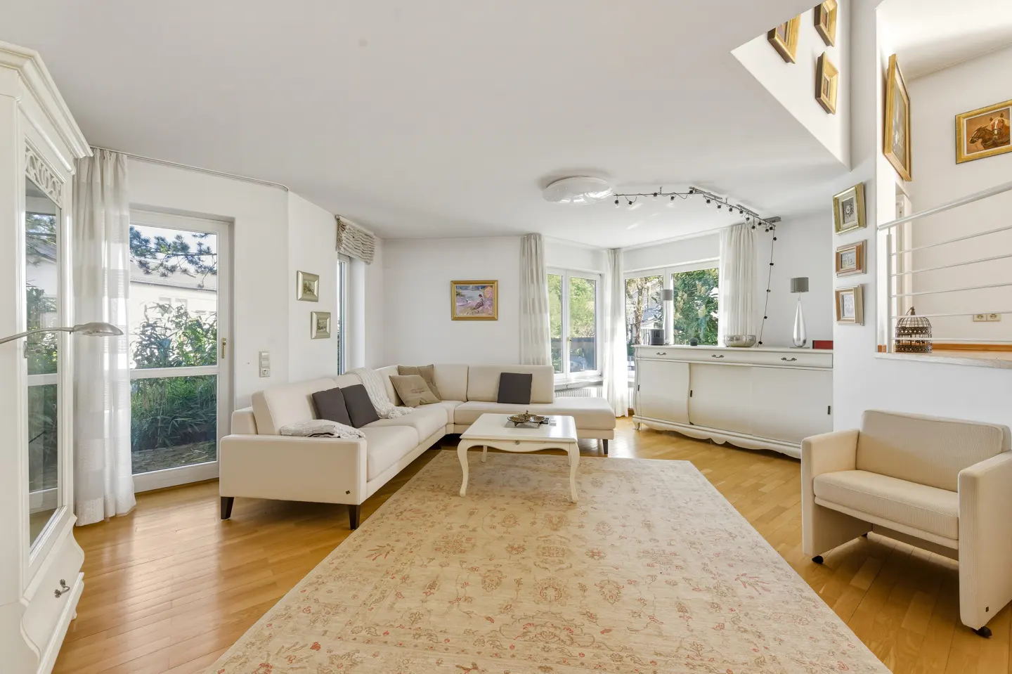 Bright living room with white walls, wood floors, and a large beige rug. A white sectional sofa and armchair provide seating. Windows offer natural light.