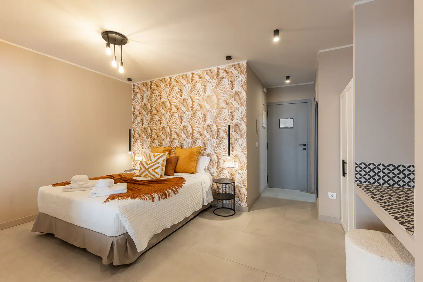 Bedroom with a white bed, orange pillows, and a patterned accent wall. A gray door and a small table are also visible.