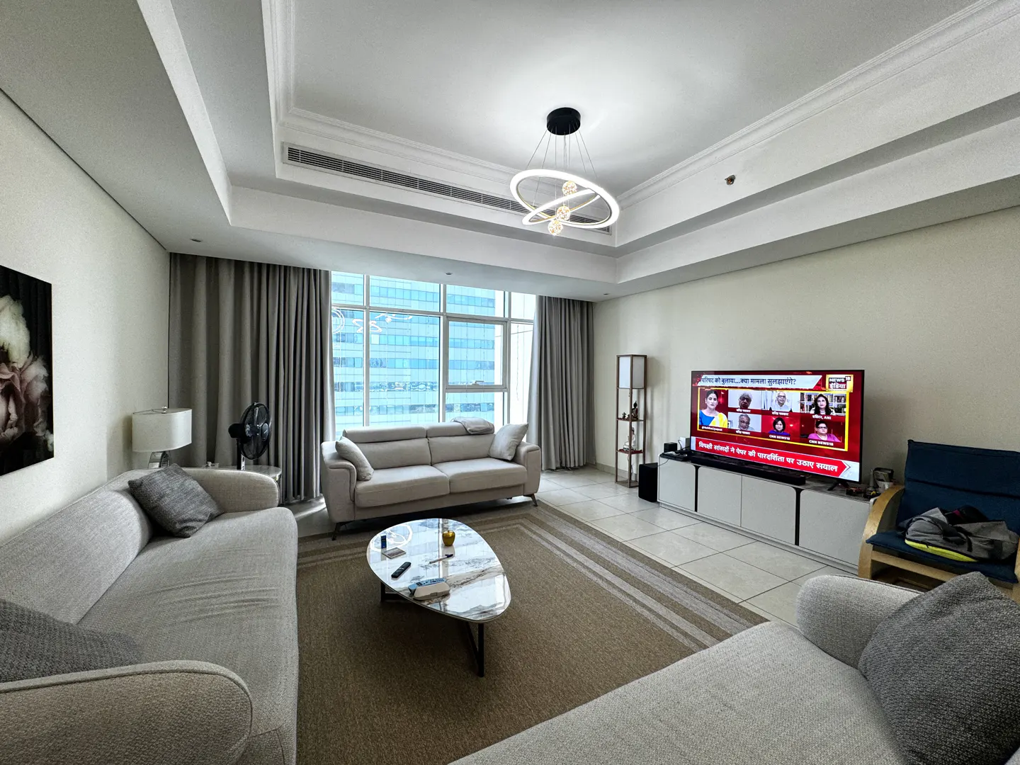 A bright living room with two gray sofas, a marble coffee table, and a large window with city views. A TV displays a news program.